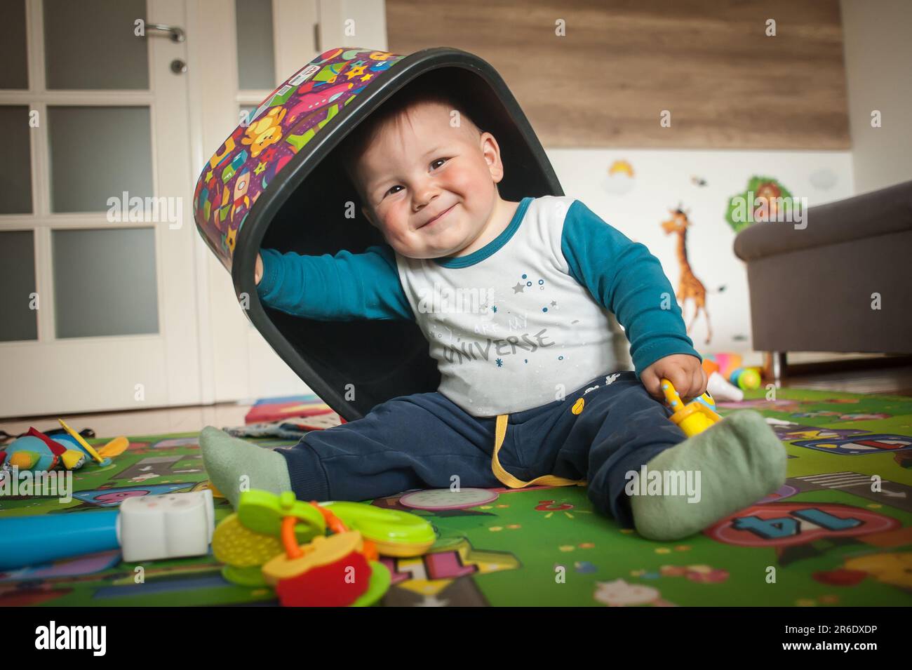 El bebé sonriente está sentado en el piso de casa, jugando con juguetes coloridos y llevando una caja de juguetes sobre su cabeza. Amor y emoción familiar Foto de stock