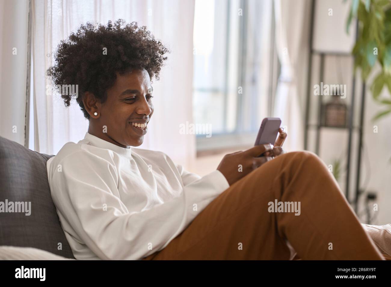 Feliz gen z adolescente afroamericano sentado en el sofá usando el teléfono en casa. Foto de stock