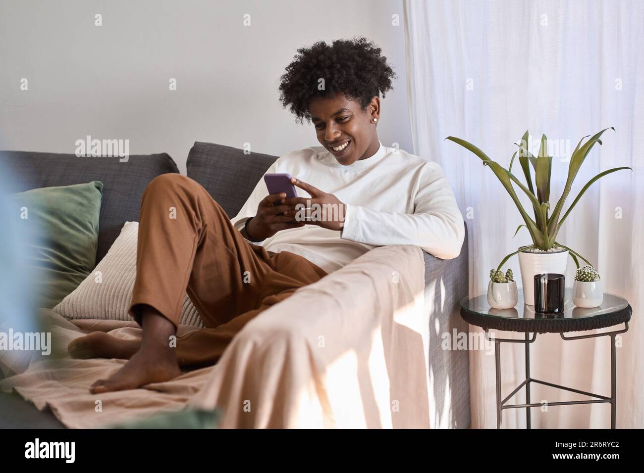 Feliz gen z adolescente afroamericano sentado en el sofá usando tecnología de teléfono móvil. Foto de stock