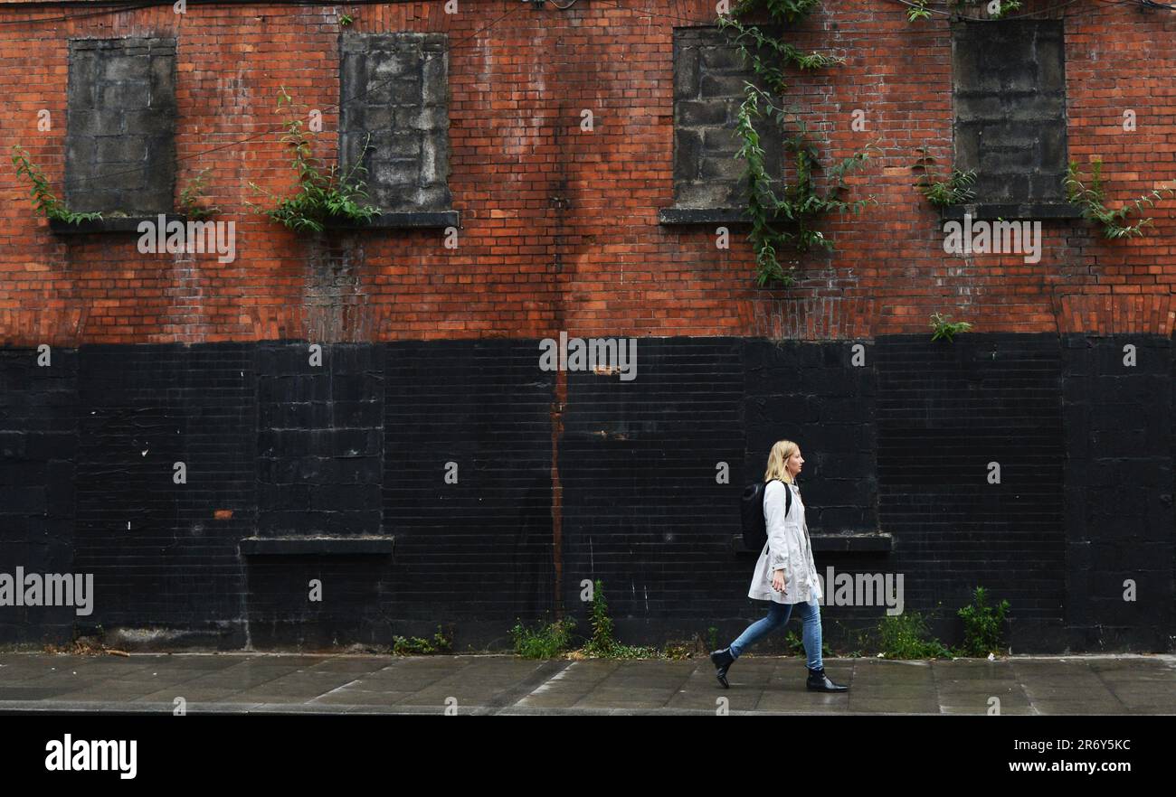 Una mujer irlandesa caminando por un antiguo edificio abandonado de fábrica de ladrillo rojo cerca del río Liffey en Dublín, Irlanda. Foto de stock