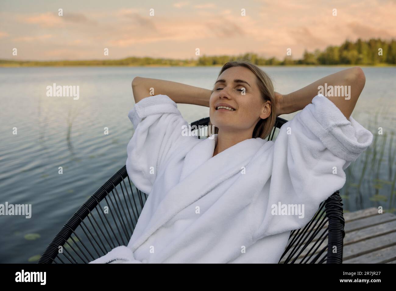 mujer en bata de baño blanca que se relaja en silla en pasarela del lago en la tarde de verano caliente. disfrute de la naturaleza Foto de stock