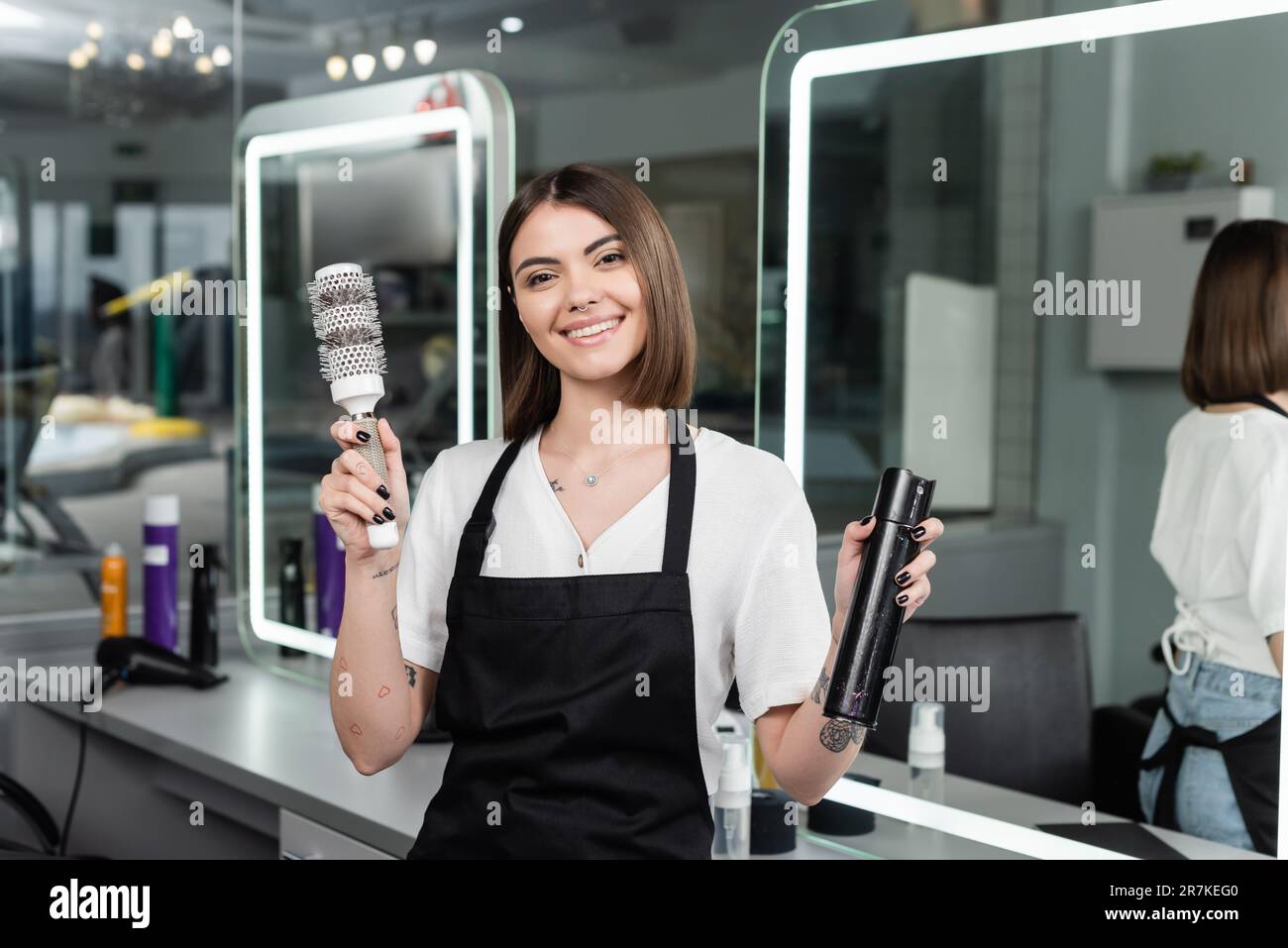 disparos de cabeza profesionales, positividad, peluquería tatuada en delantal sosteniendo cepillo redondo y botella con spray para el cabello, trabajador de belleza, mirando a la cámara, sm Foto de stock