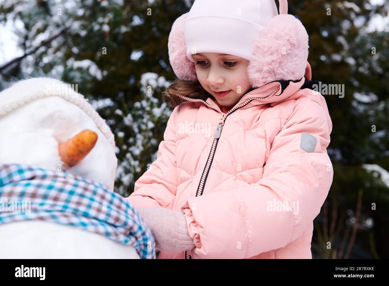 Closeup niña encantadora en chaqueta rosa abajo y orejeras esponjosas,  poniéndose una bufanda azul alrededor del cuello del muñeco de nieve  Fotografía de stock - Alamy