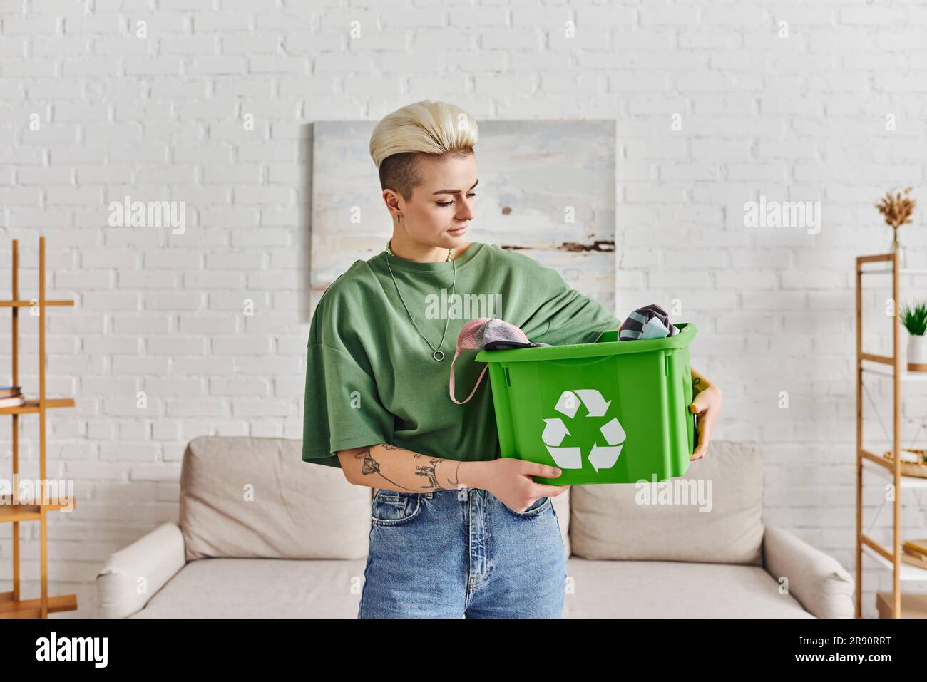 estilo de vida eco-consciente, mujer joven y tatuada con peinado de moda que sostiene la caja de reciclaje verde con la ropa, la vida sostenible y el ecologismo Foto de stock