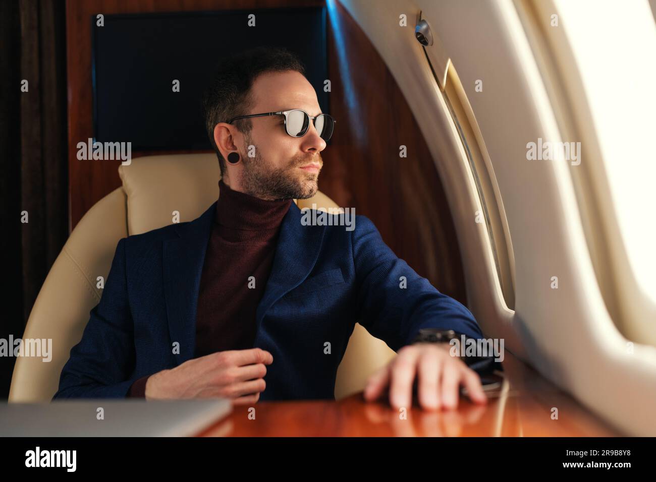 Elegante CEO confiado Hombre de negocios con gafas de sol en una chaqueta azul que se sienta dentro de un avión privado de primera clase que mira a través de una ventana Foto de stock