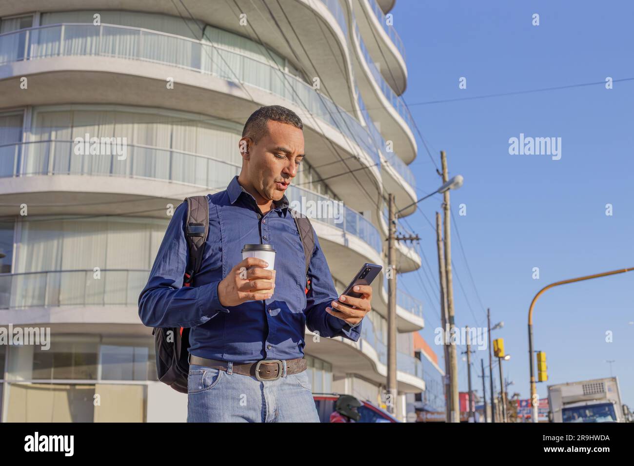 Joven latino mirando su teléfono móvil con cara sorprendida y con una taza de papel con café en la mano. Foto de stock