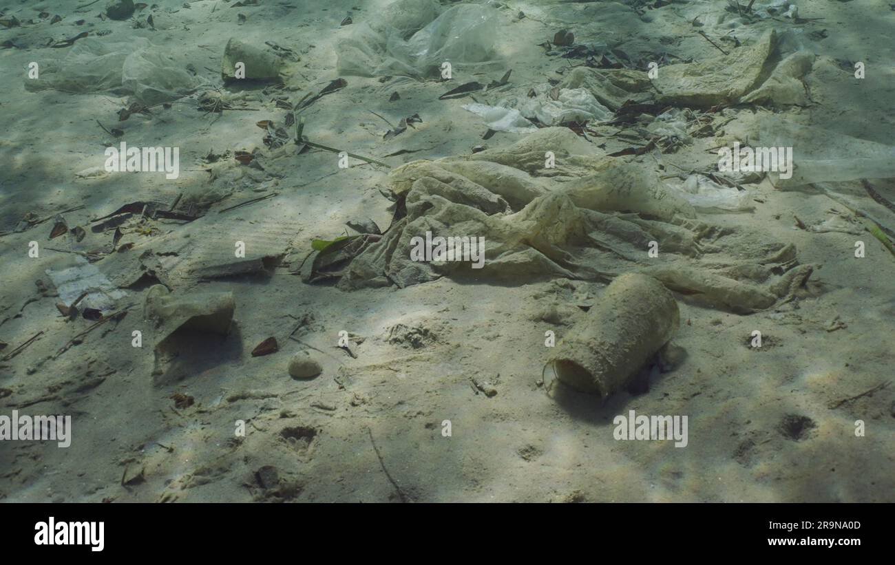 La basura plástica se encuentra en el fondo arenoso-limo del mar en aguas poco profundas en el día soleado brillante en el resplandor del sol. Fondo marino contaminado con plástico y otros residuos, Re Foto de stock