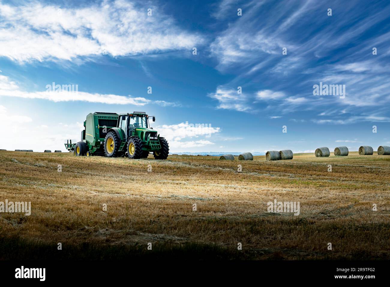 Un tractor que tira de una empacadora a través de un campo cosechado con balas de paja redondas en el fondo bajo un cielo azul profundo en el condado de Rocky View, Alberta, Canadá. Foto de stock