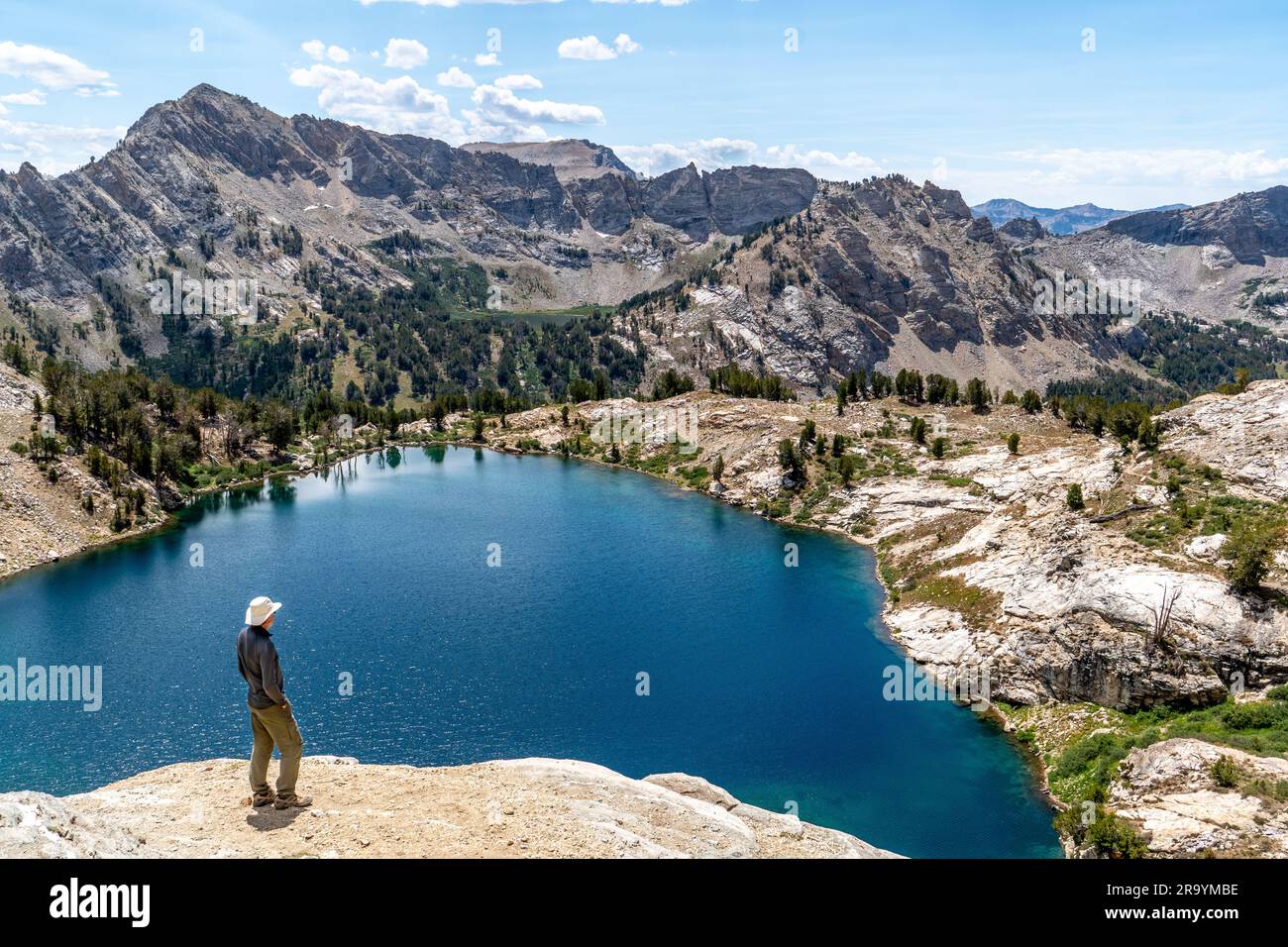 Hombre, excursionista, de pie, viendo una escena de un lago alpino y montañas escarpadas con árboles, rocas y nubes hinchadas en el cielo azul, Liberty Lake, Ruby Foto de stock
