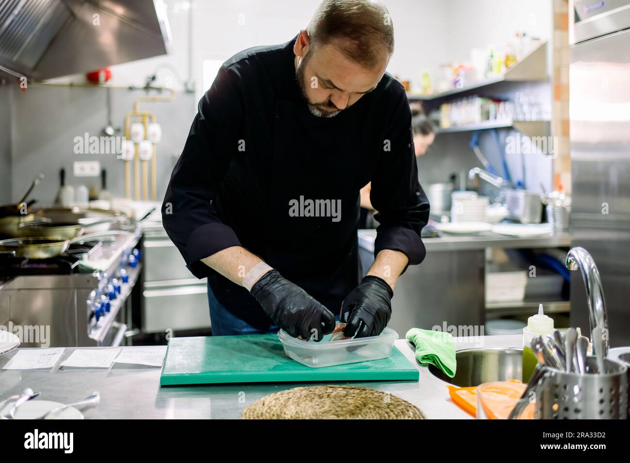 Gruñón etc. Privación Chef profesional masculino barbudo en guantes negros que arraigan la comida  en un recipiente transparente mientras trabaja en la cocina profesional  Fotografía de stock - Alamy