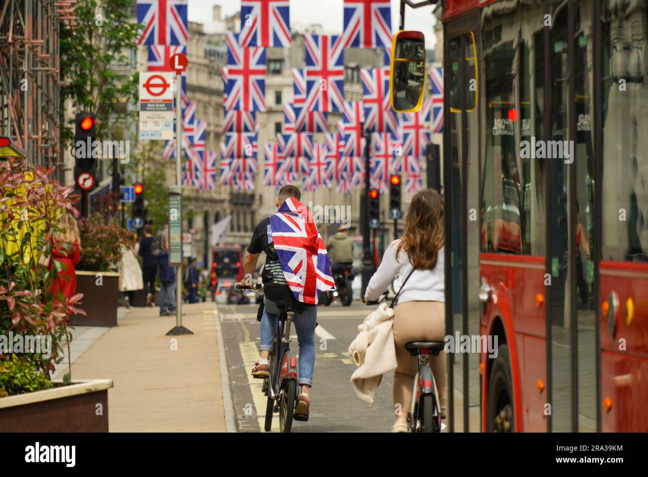 Un hombre envuelto en una bandera de Union Jack muestra su orgullo mientras monta una bicicleta en la bandera del Reino Unido bordeada por Regent Street, llena de autobuses durante la Semana de la Coronación. Foto de stock