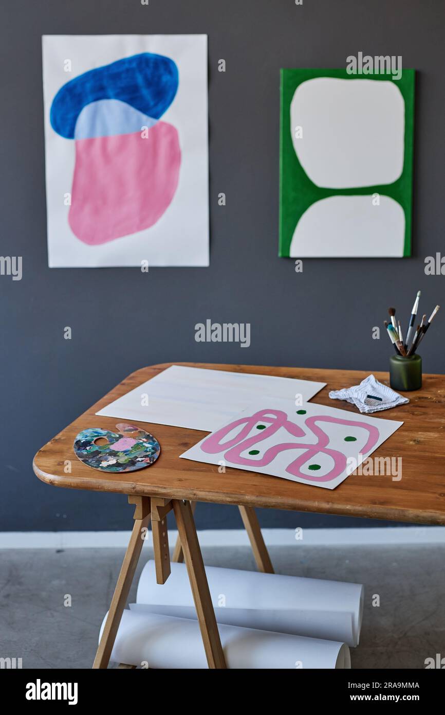 Escritorio de madera con hojas de papel, paleta de acuarela y pequeño tarro con pinceles de pie contra la pared gris con obras de arte creativas Foto de stock