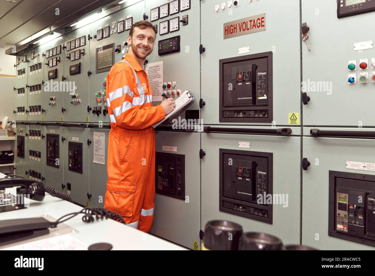 Joven oficial de ingeniería inspeccionando la centralita eléctrica en la sala de control del motor. Ingeniero offshore sonriendo. Foto de stock