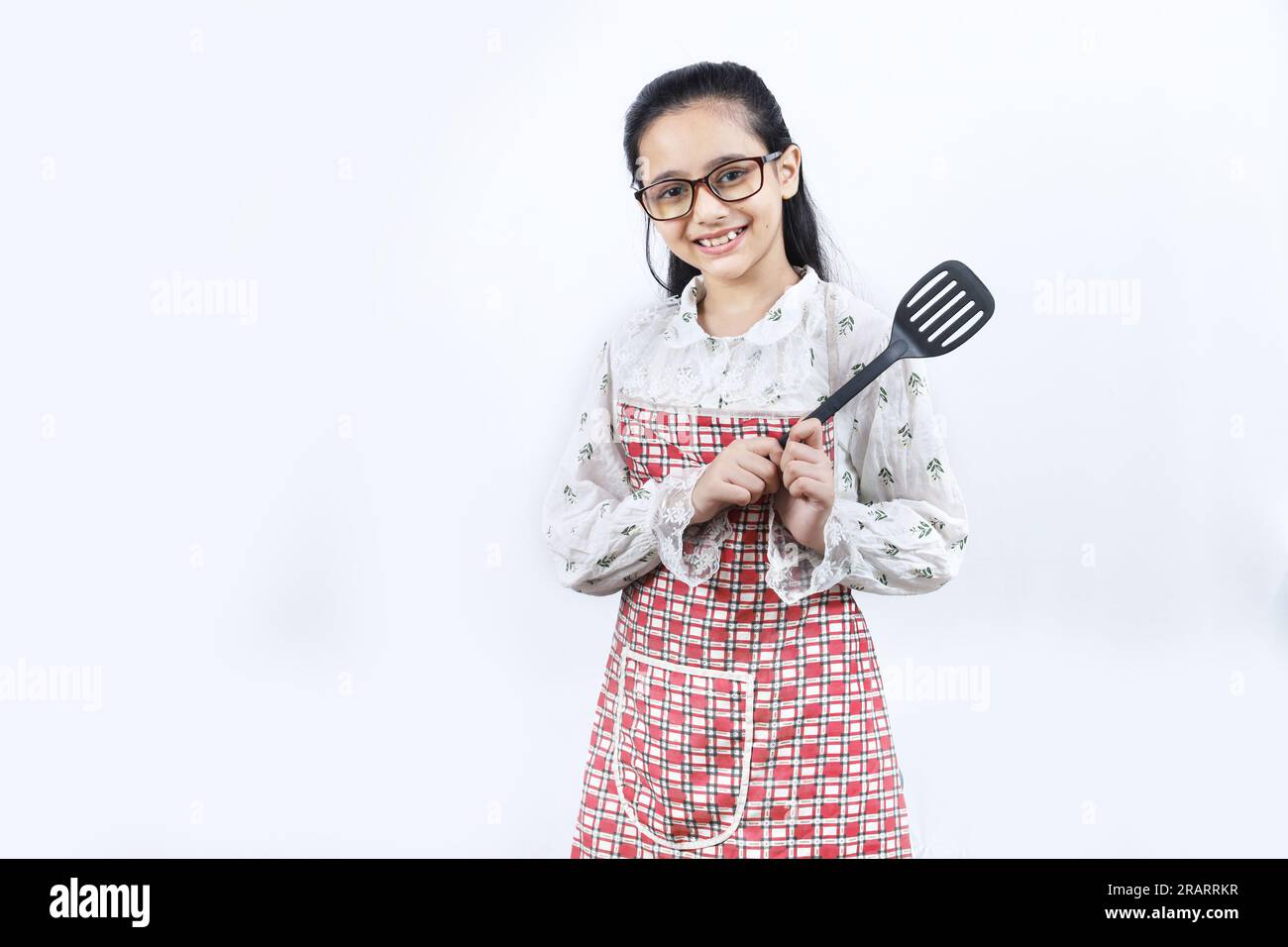 Retrato de la muchacha adolescente india feliz en la cocina que sostiene los accesorios de cocina. Rin enrollable, cuchara de freír, cucharón, sartén. Joven alegre y alegre Foto de stock