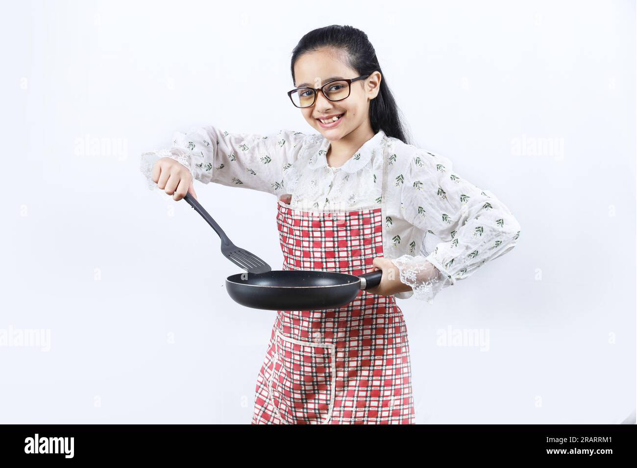 Retrato de la muchacha adolescente india feliz en la cocina que sostiene los accesorios de cocina. Rin enrollable, cuchara de freír, cucharón, sartén. Joven alegre y alegre Foto de stock