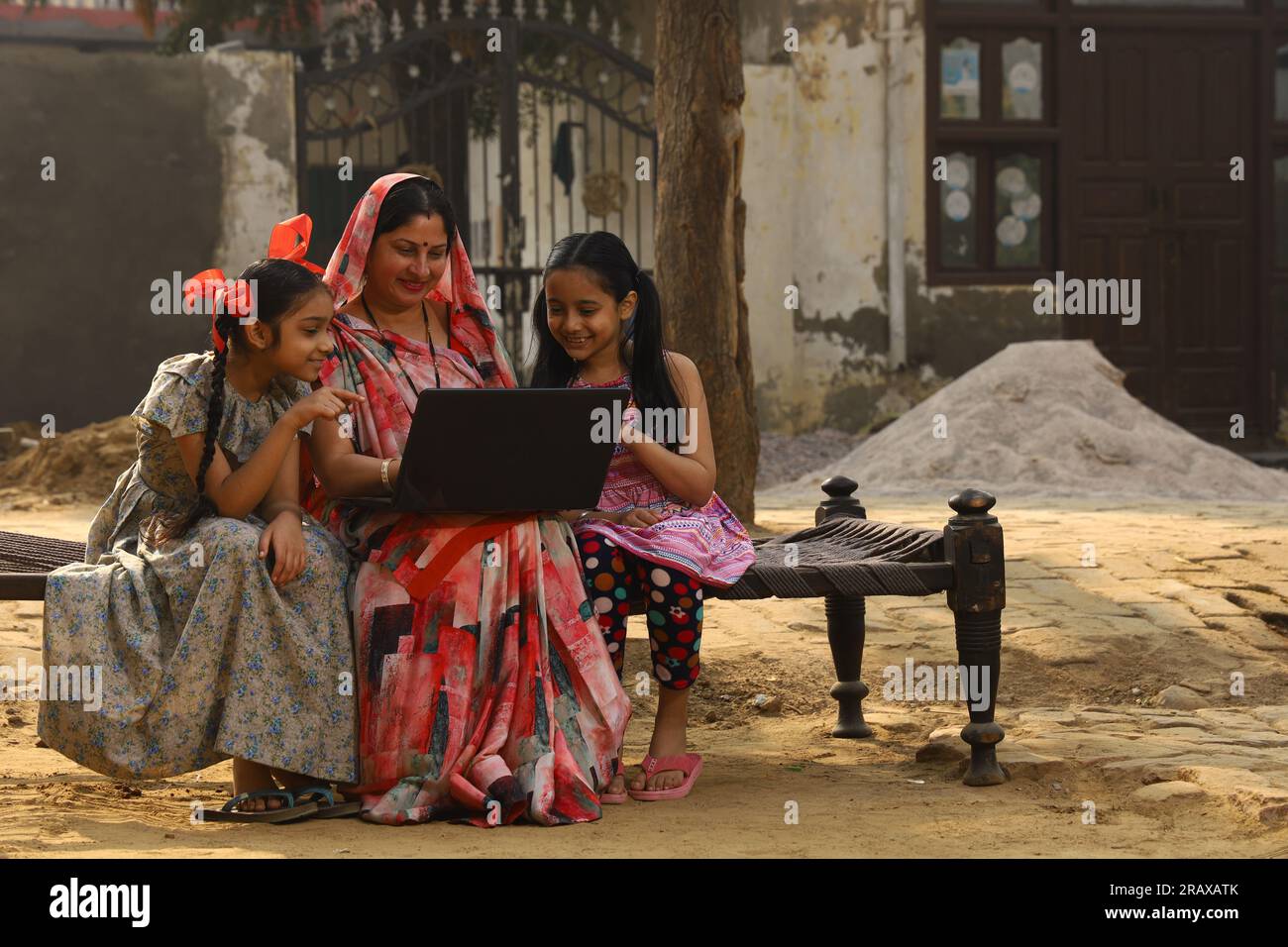 Familia rural india feliz en el pueblo. Madre e hijas sentadas juntas sonriendo en la cuna fuera de su casa en el patio delantero. Madre en hermoso sari Foto de stock