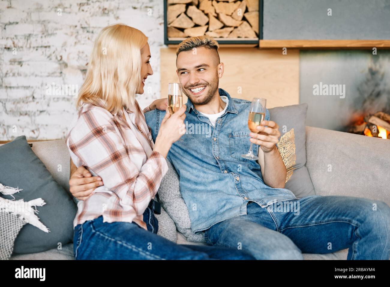 Joven pareja feliz bebiendo champán y sonriendo sentado en el sofá en su casa moderna. Relación, celebración, concepto de citas Foto de stock