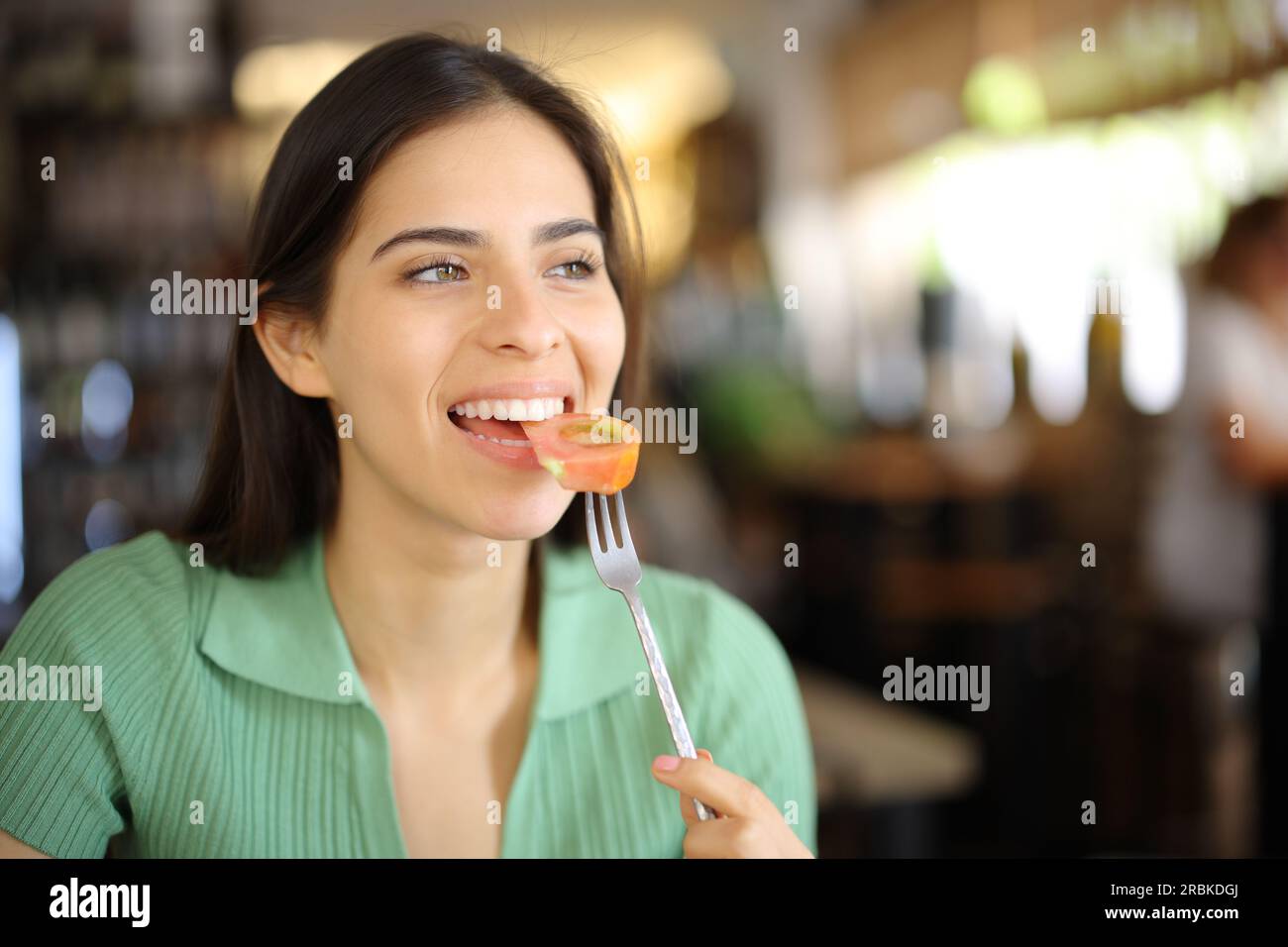Mujer feliz comiendo tomate en el interior de un restaurante mirando lejos Foto de stock