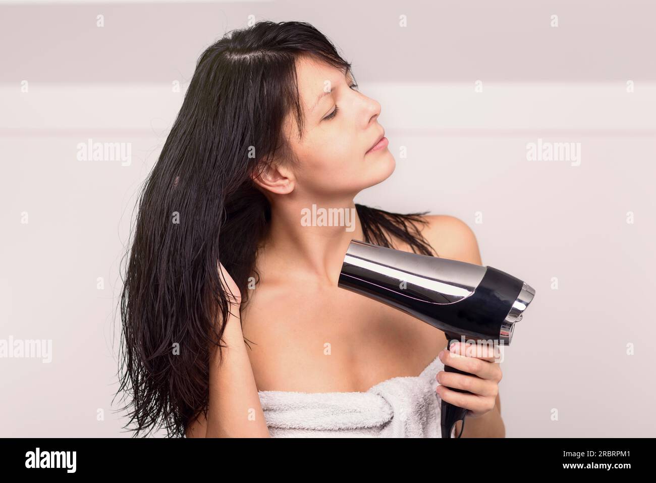 Mujer atractiva en un soplado de toalla que seca su pelo castaño de tronco con un secador de pelo de mano en su cuarto de baño mirando a la cámara con una sonrisa amistosa cálida Foto de stock