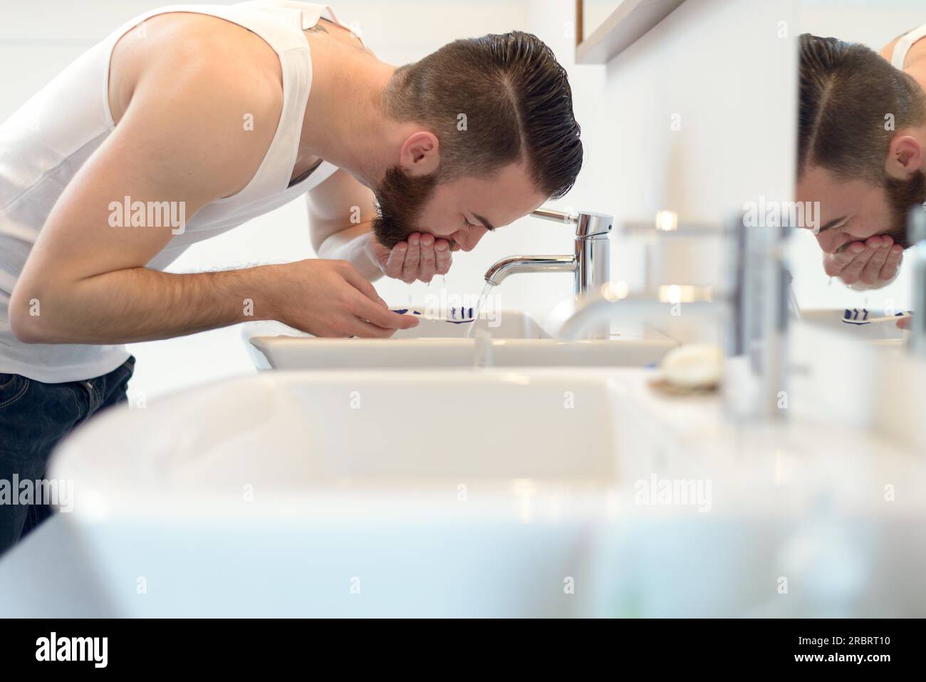 Hombre enjuagando su cepillo de dientes con agua corriente del grifo en el lavabo después de afeitarse por encima de su barba para limpiar su apariencia, vista lateral Foto de stock
