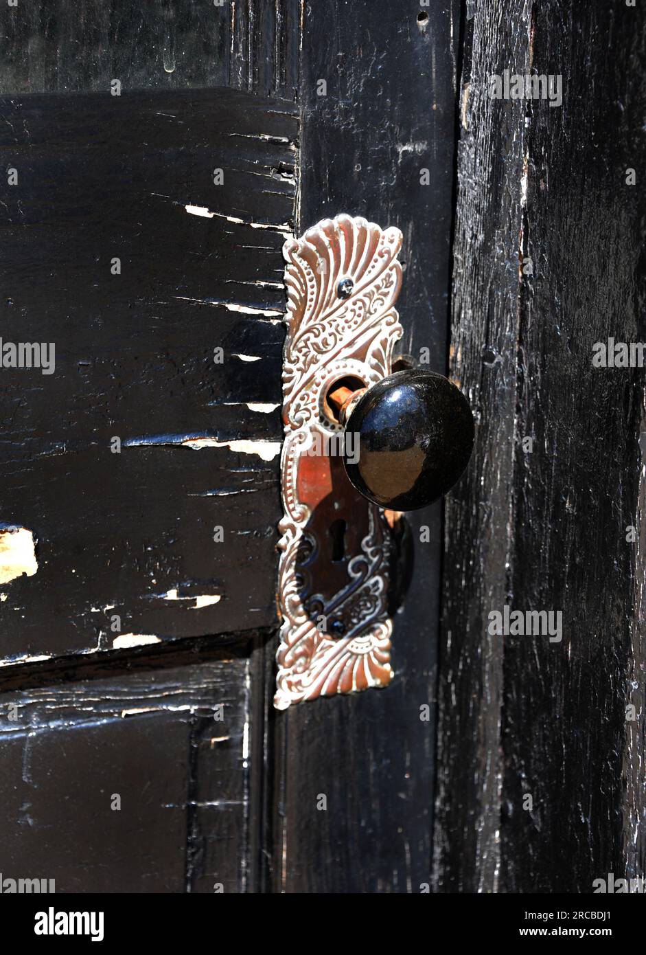 La puerta de madera deteriorada es de color negro trenzado. La pintura se está pelando y la madera está agrietada. La placa de la puerta antigua y la perilla están fijadas a la puerta. Diseño intrincado Foto de stock