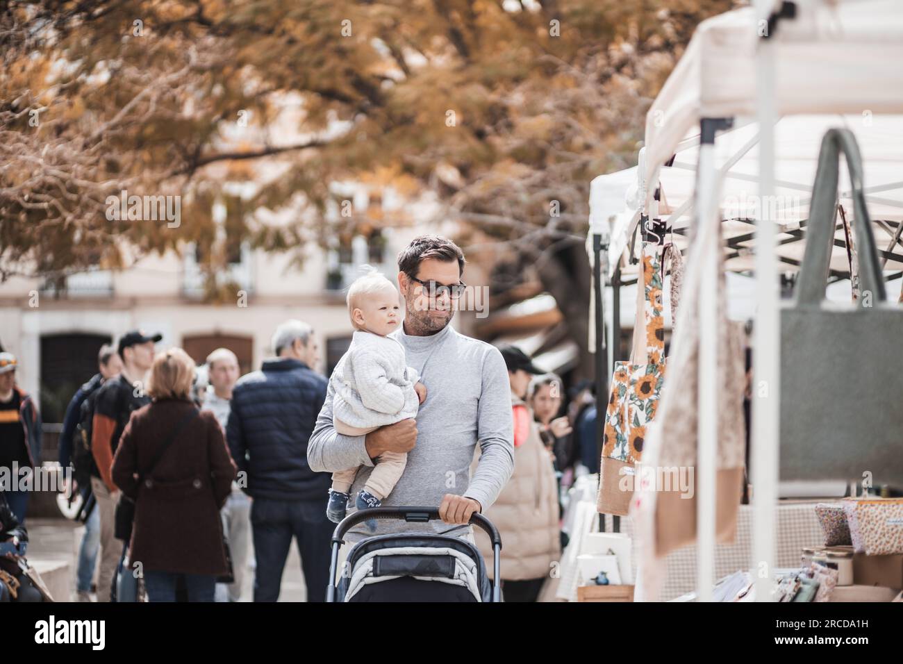 Padre caminando llevando a su bebé niño y empujando el cochecito en la multitud de personas que vigila el mercado de pulgas del domingo en Málaga, España Foto de stock
