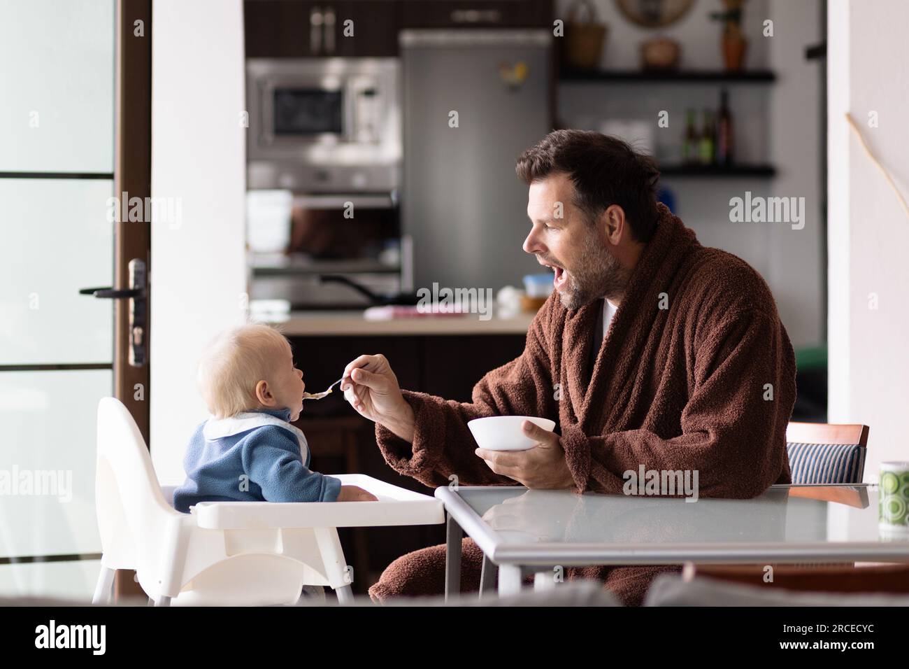 Padre con cuchara de cuerda de baño alimentación hir niño bebé niño sentado en silla alta en la mesa de comedor en la cocina en casa por la mañana. Foto de stock