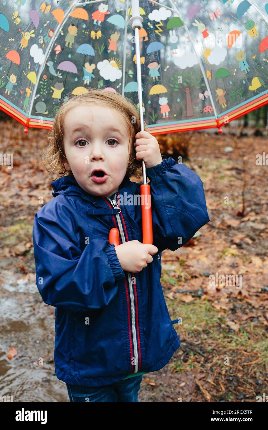 Niño pequeño sosteniendo paraguas bajo la lluvia y mirando sorprendido Foto de stock