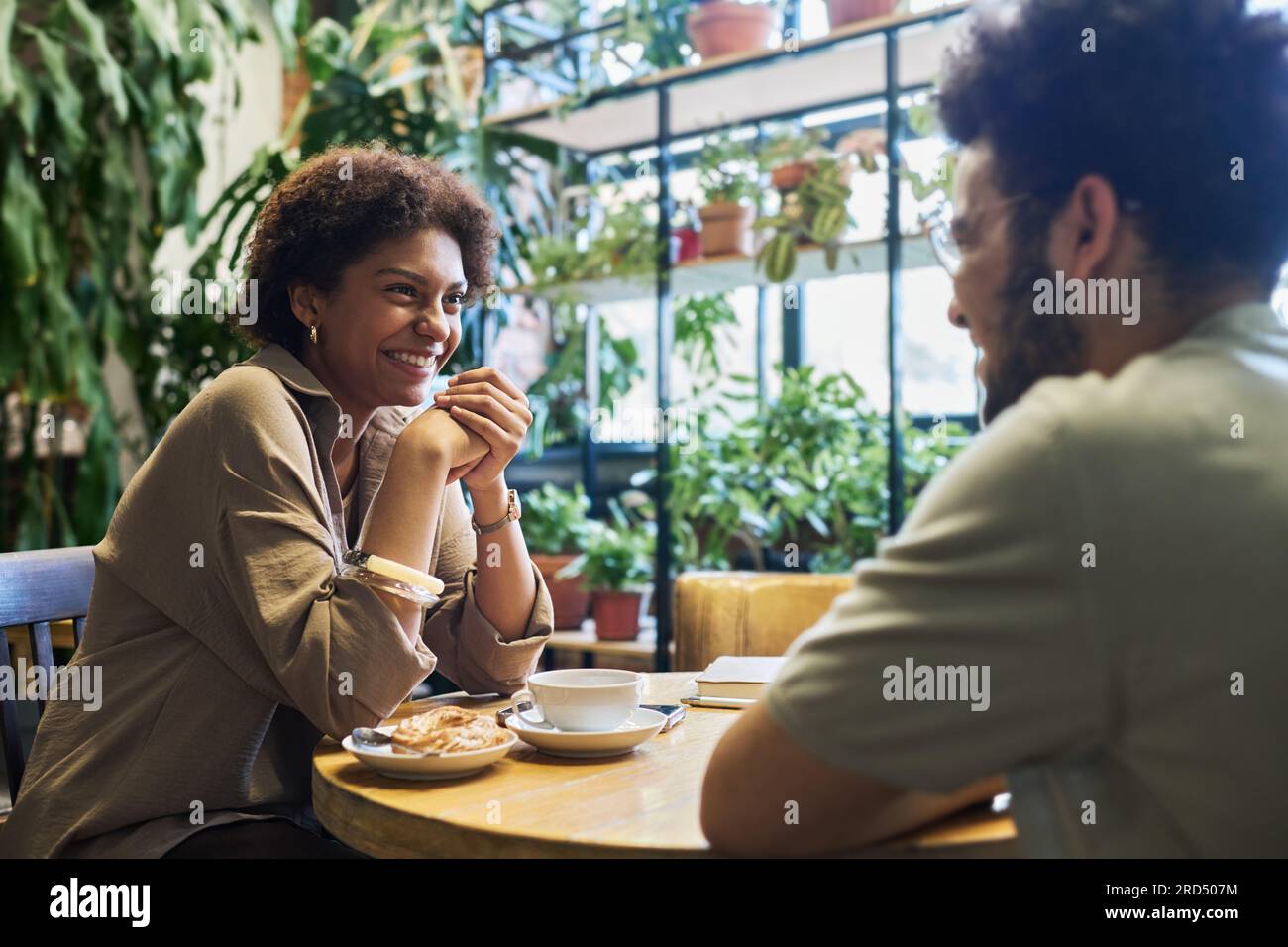 Mujer joven feliz mirando a su novio con sonrisa dentada mientras ambos se sientan uno frente al otro en la mesa y disfrutando de la fecha en el café Foto de stock
