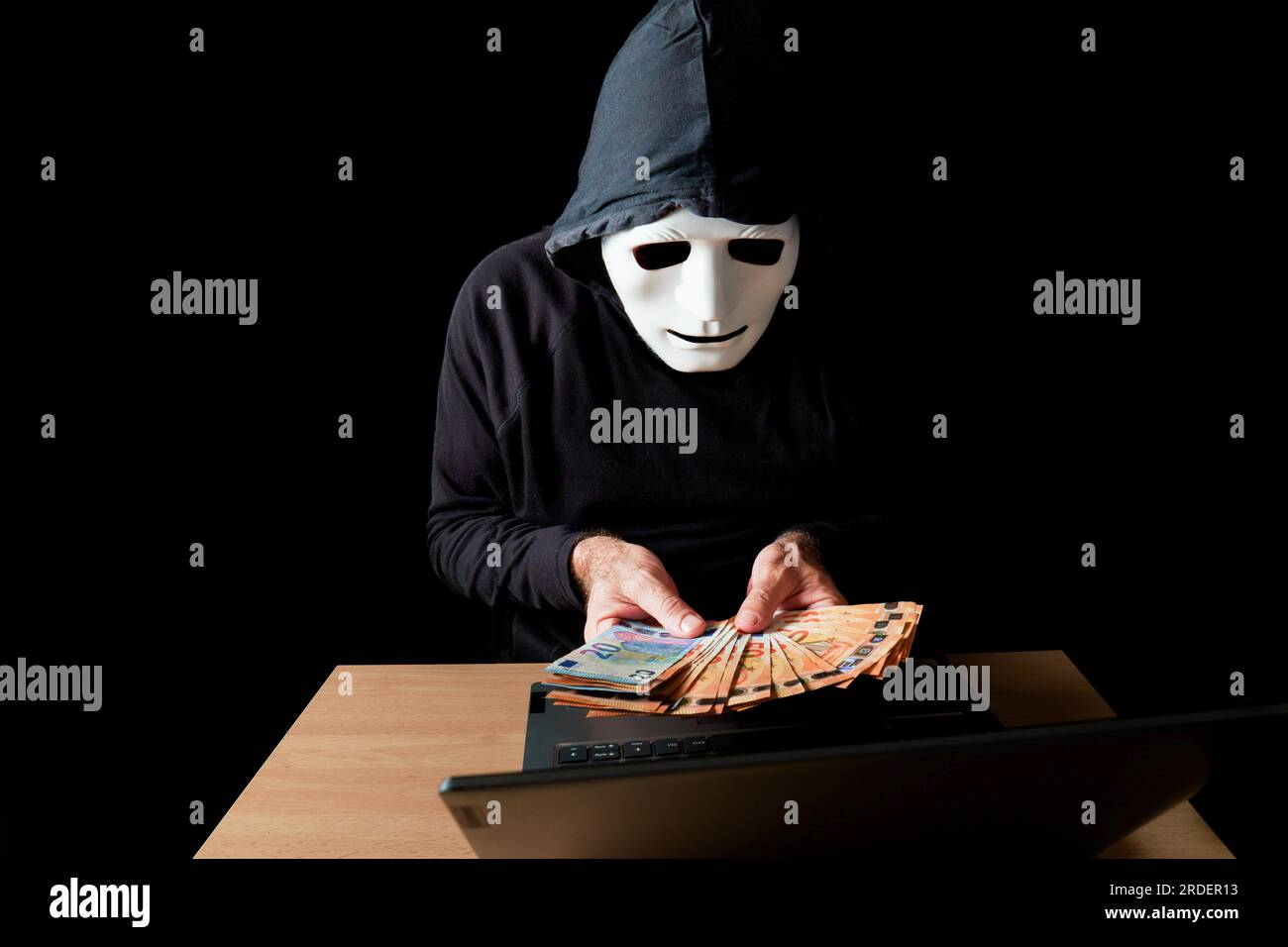 Hombre hacker vestido de negro con máscara blanca y capucha negra contando billetes de euro en sus manos, concepto de cibercrimen, aislado sobre fondo negro Foto de stock