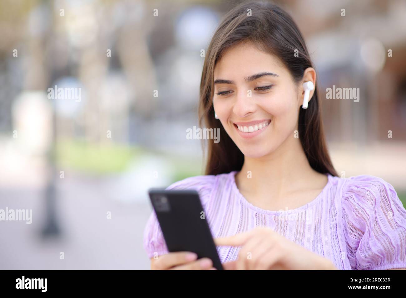 Mujer feliz con el auricular y el teléfono caminando en la calle Foto de stock