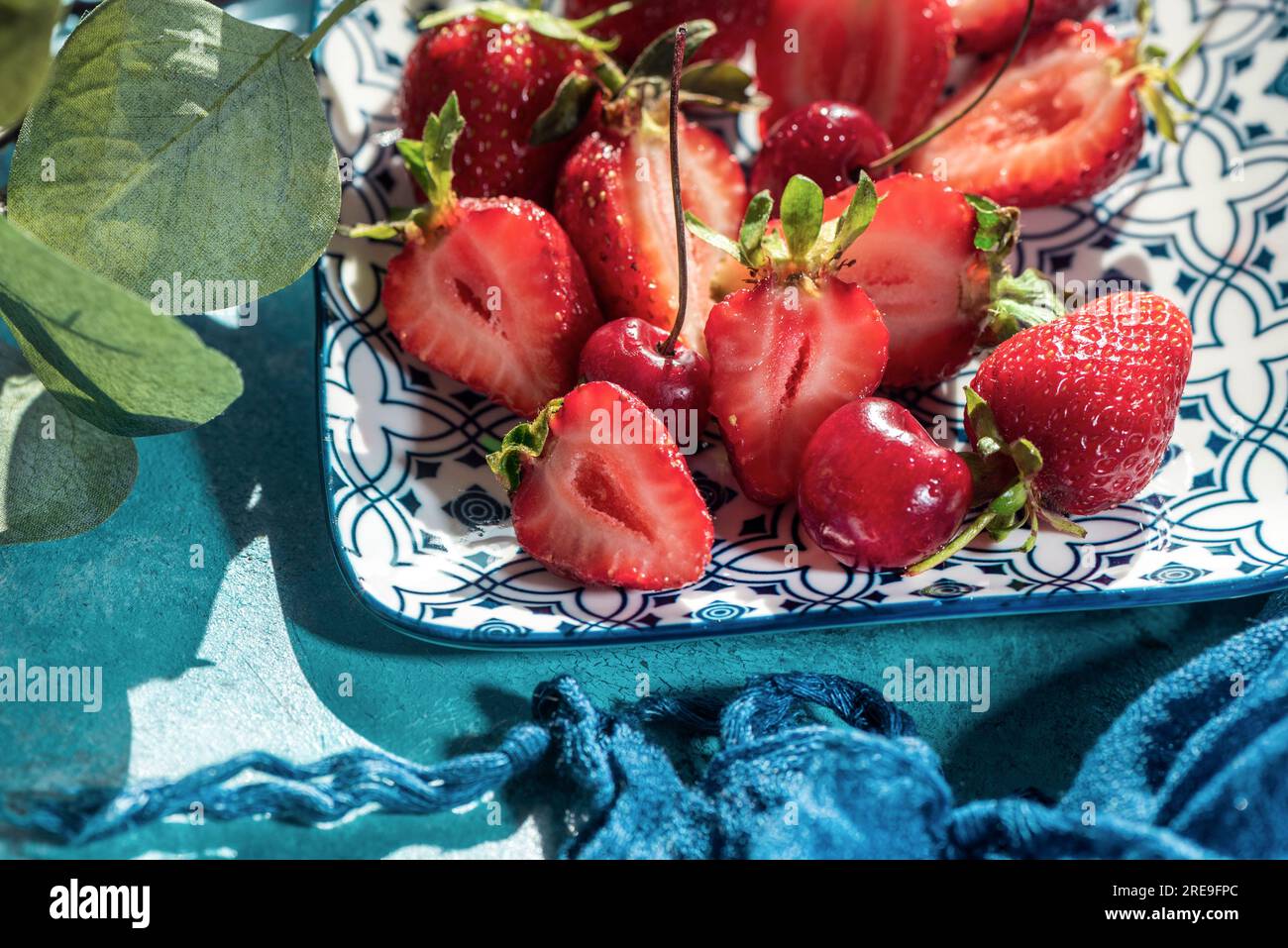 Frutas rojas frescas en un plato de cerámica con plantas sobre un fondo de madera azul, cerca, fotografía de naturaleza muerta Foto de stock