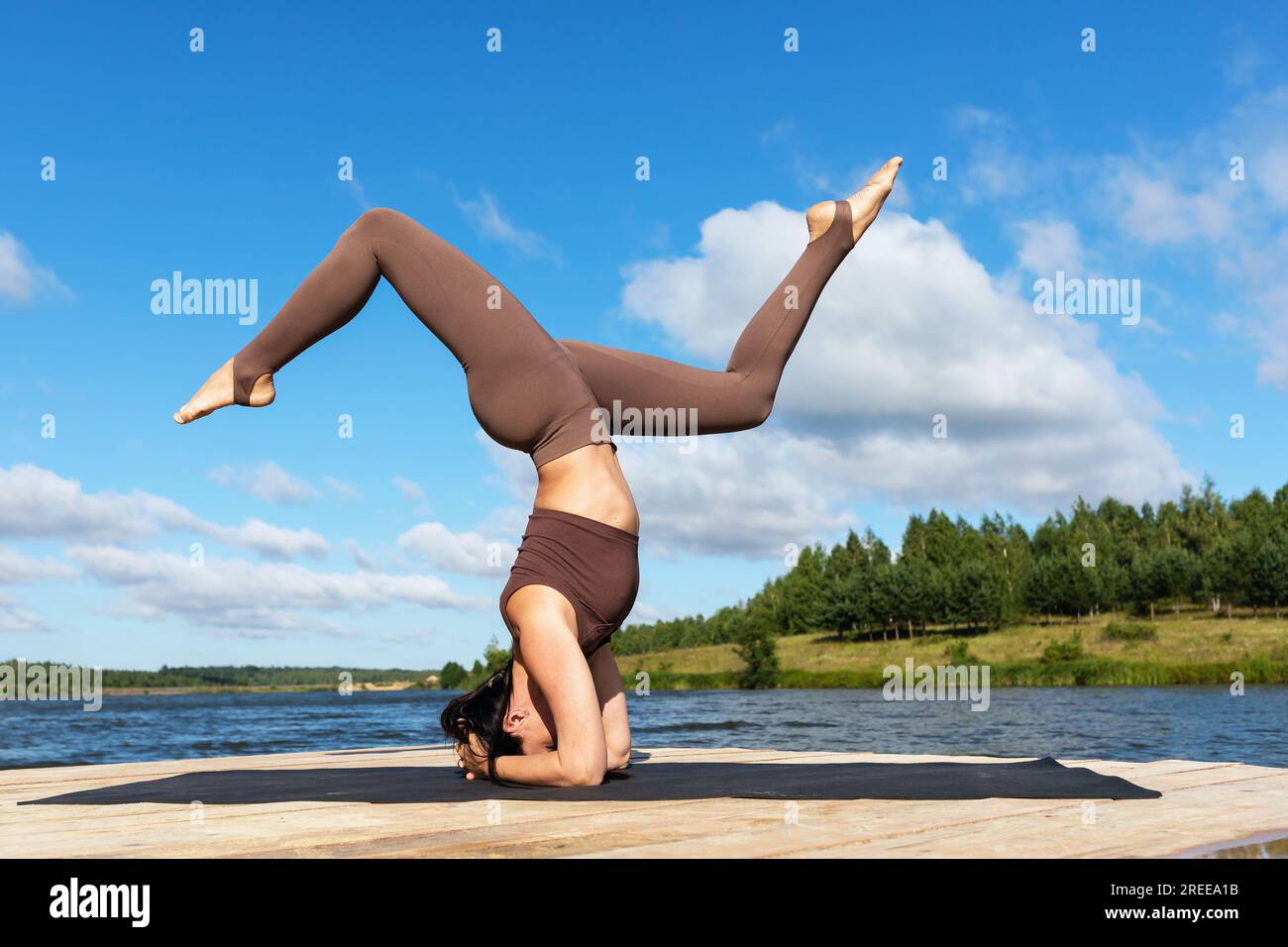 Una mujer practicando yoga, realiza una variación del ejercicio Shirshasana, asana invertida, pie de cabeza y pie de mano, entrena en ropa deportiva en la orilla Foto de stock