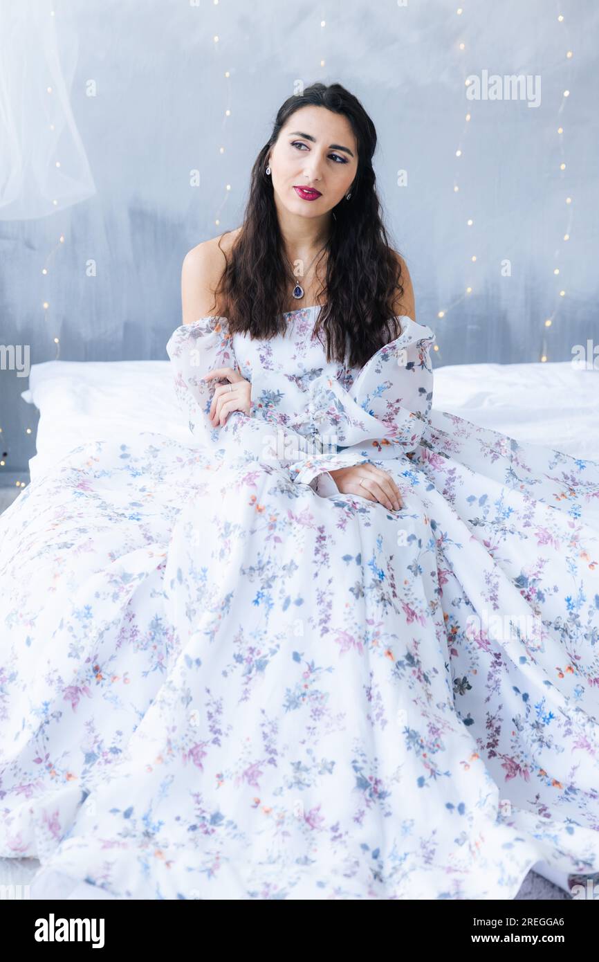 Una chica en una cama con un vestido blanco mira hacia otro lado sobre un fondo gris Foto de stock