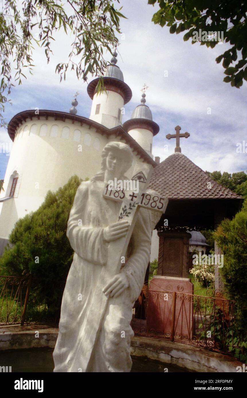 Monasterio de Varatec, Condado de Neamt, Rumania, 1999. Una estatua de un ángel sosteniendo una cruz en los terrenos de la primera iglesia de madera del monasterio (construido en 1785). En la parte posterior, la iglesia 'Dormición de la Virgen María'. Foto de stock