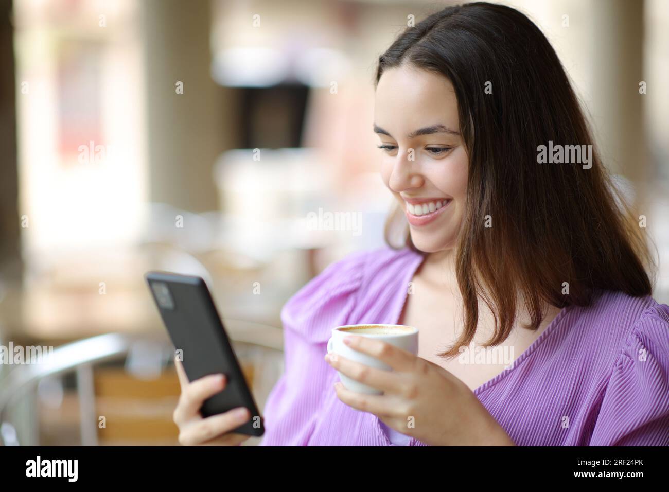 Mujer feliz comprobando el teléfono celular y sonriendo en una terraza de cafetería Foto de stock