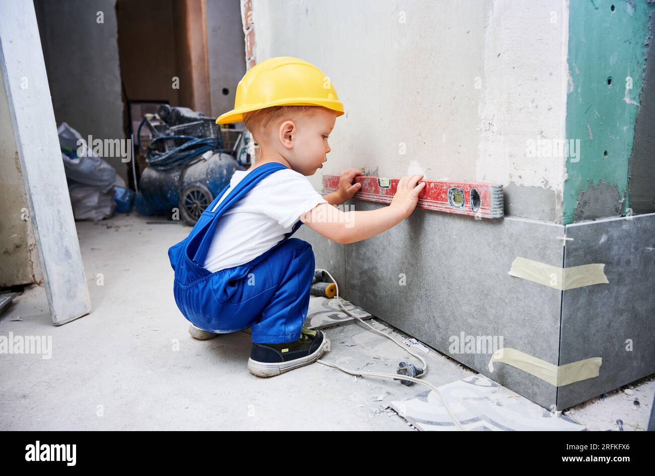 Pequeño trabajador de la construcción del muchacho que mide la superficie  de la pared con la herramienta del nivel del espíritu. Niño en mono de  trabajo usando instrumento de nivel de burbuja