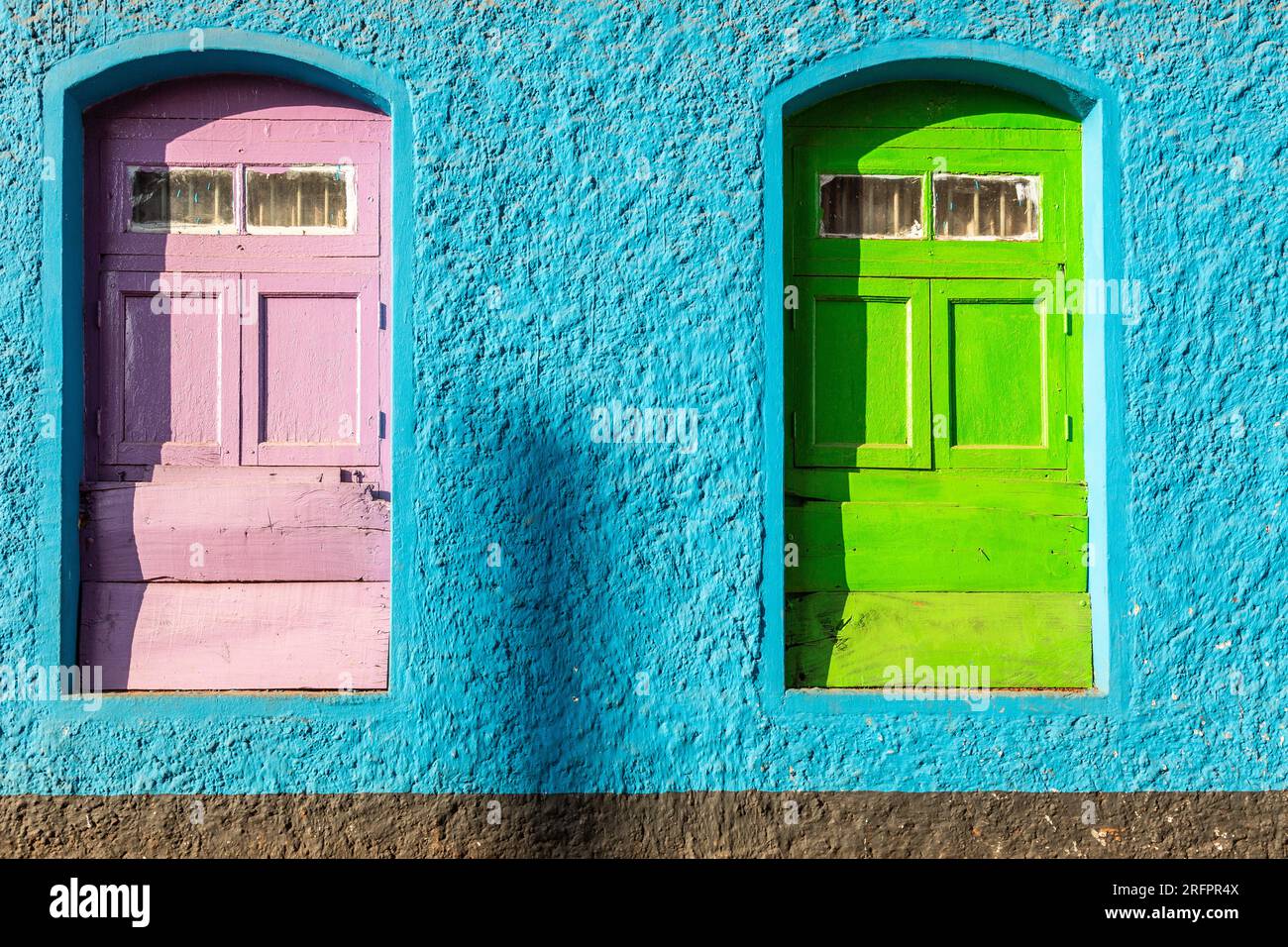Dos ventanas francesas, una rosa, la otra verde, en una pared azul. Colores llamativos. Foto de stock