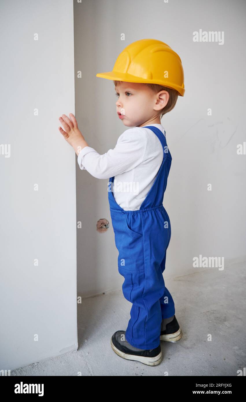 Trabajador de la construcción infantil de pie cerca de la pared en el  apartamento bajo renovación. Niño en casco de seguridad y mono de trabajo  jugando en el futuro hogar Fotografía de