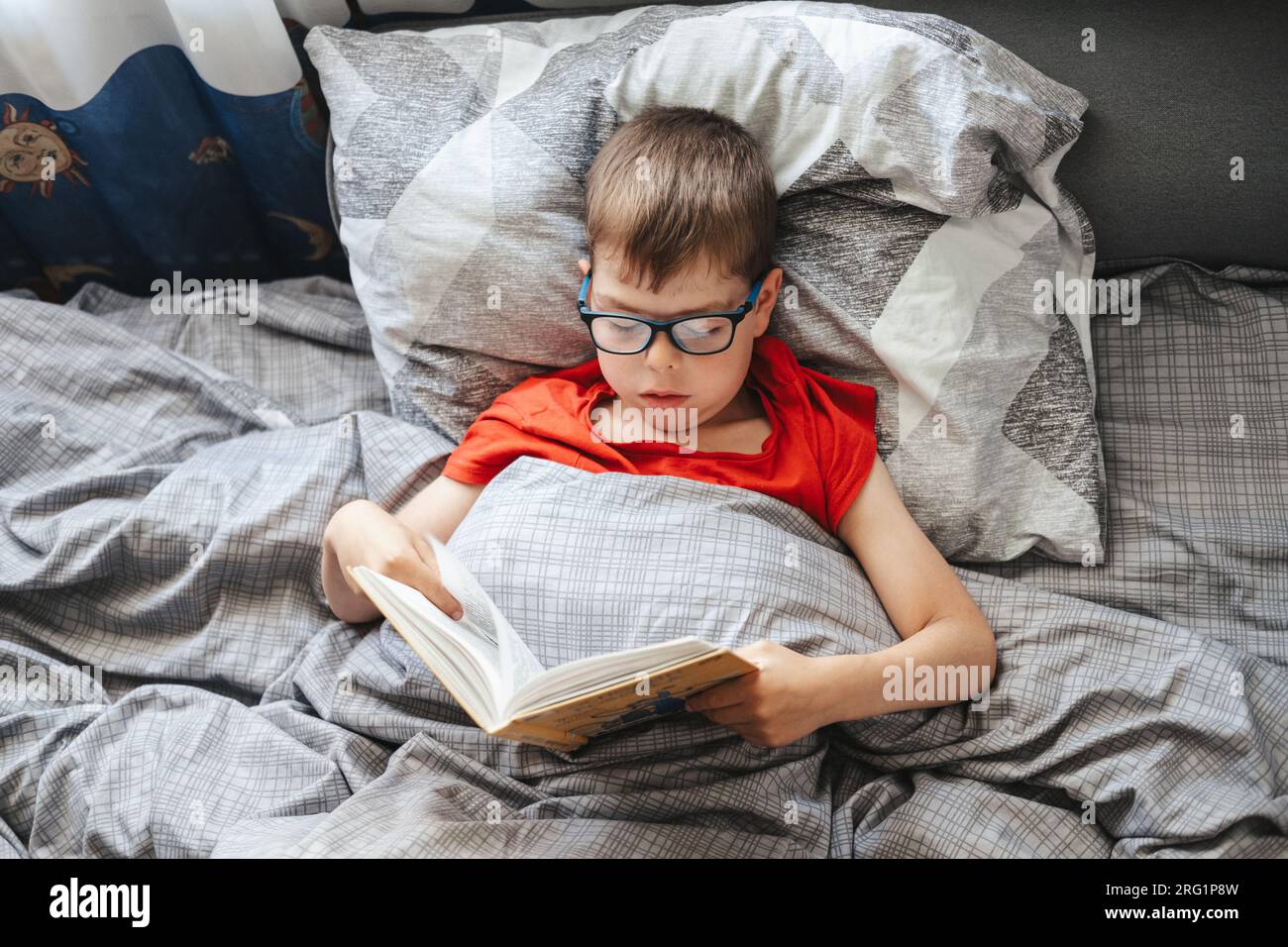 chico con gafas se encuentra en una cama debajo de una manta y lee un libro, vista superior. Un niño en edad de escuela primaria está leyendo un libro en su cama. Foto de stock
