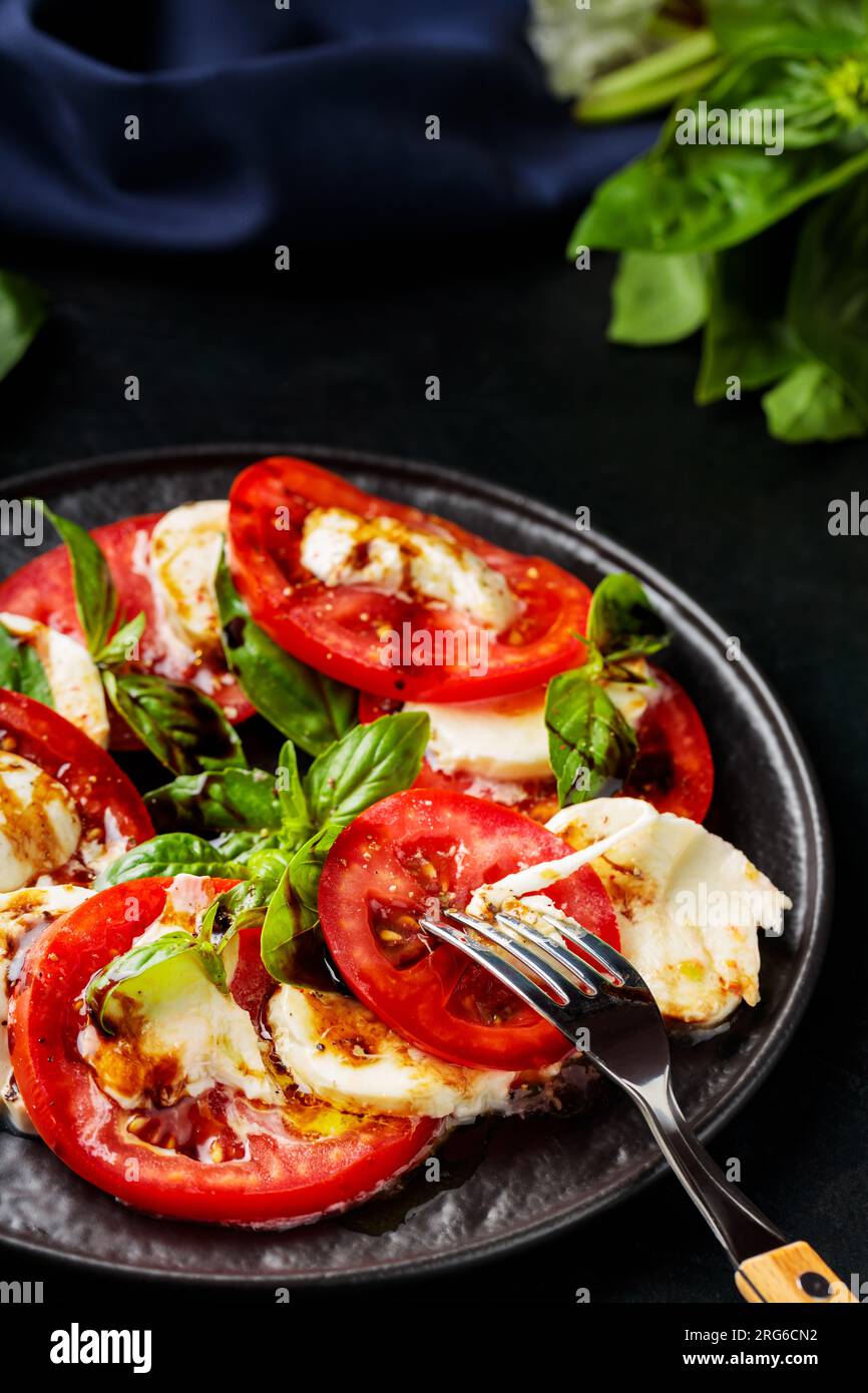 Ensalada Caprese. Rodajas de tomate y mozzarella con hojas de albahaca con tenedor en plato oscuro. Comida tradicional italiana Foto de stock