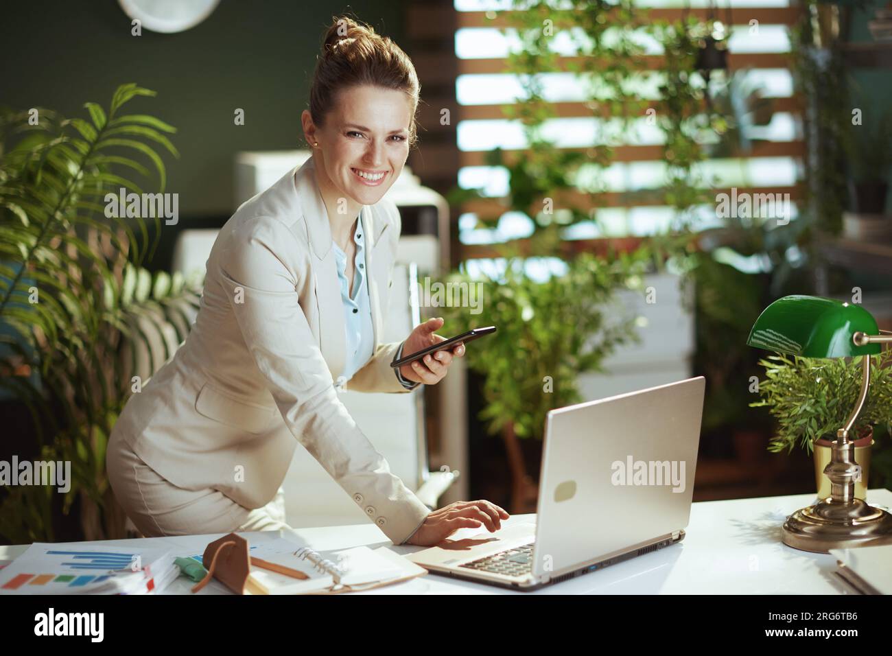 Retrato de sonriente mujer moderna de 40 años de edad contable en un traje de negocios ligero en la oficina verde moderna con el ordenador portátil usando la aplicación del smartphone. Foto de stock