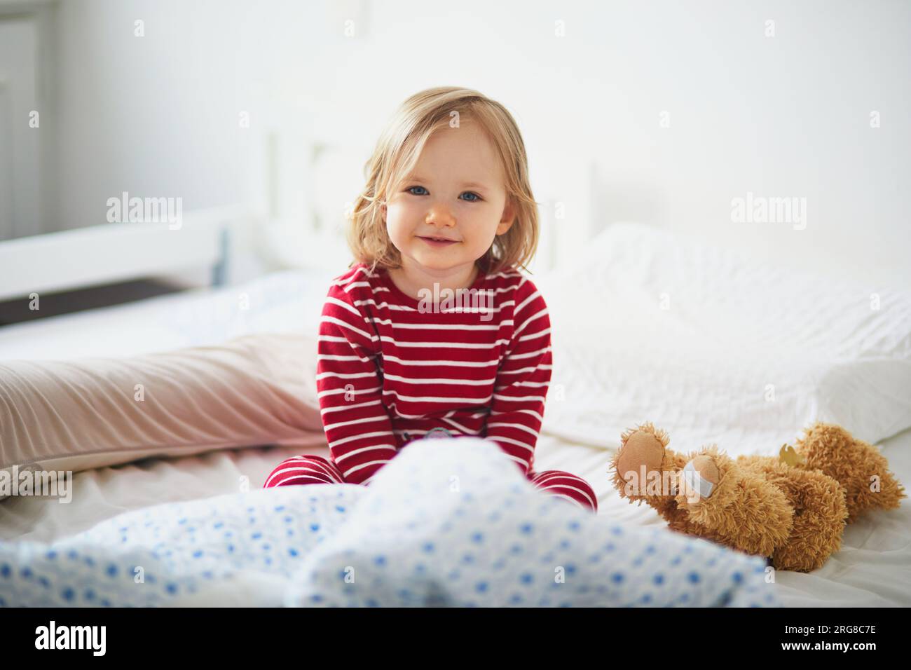 Niña feliz en pijama de rayas rojas y blancas sentado en la cama justo después de despertar. Siestas de día para niños pequeños Foto de stock