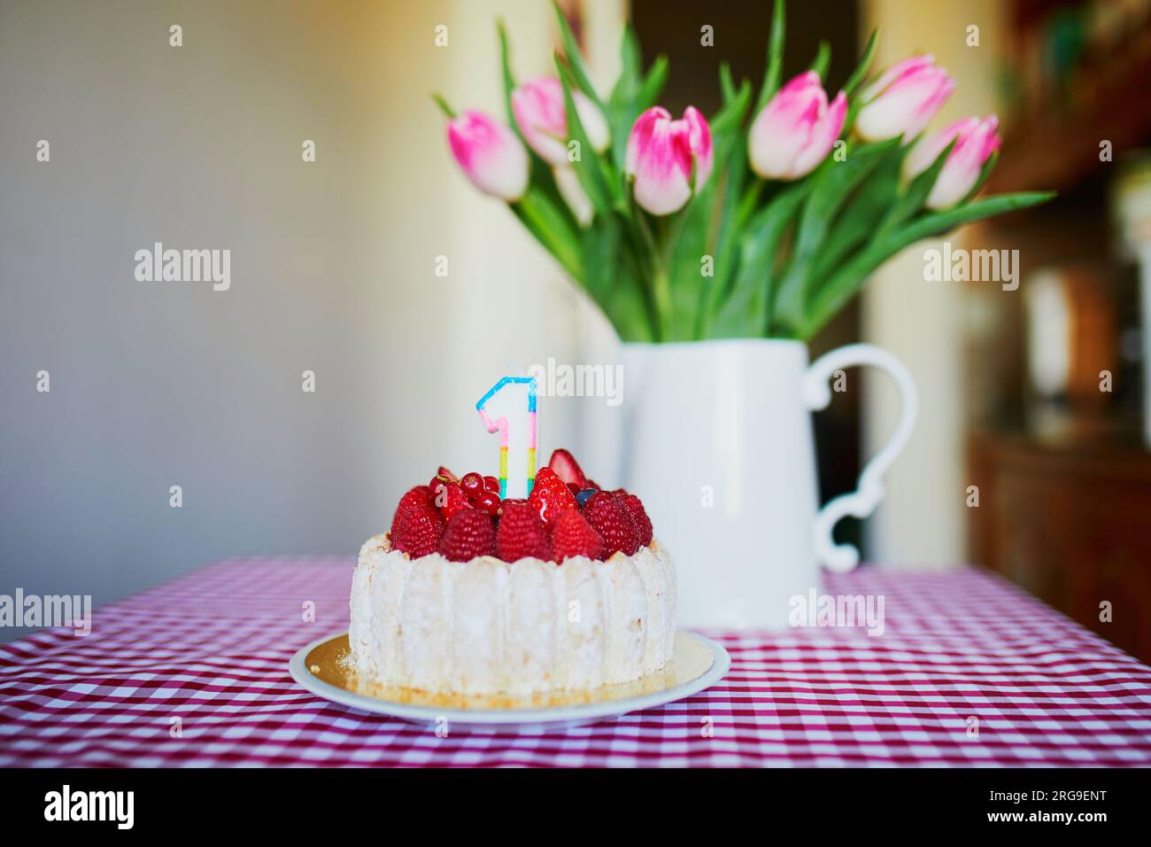Delicioso pastel de frutas con frambuesas y vela en forma de número uno con un montón de hermosos tulipes rosas en jarrón. El primer cumpleaños del bebé Foto de stock