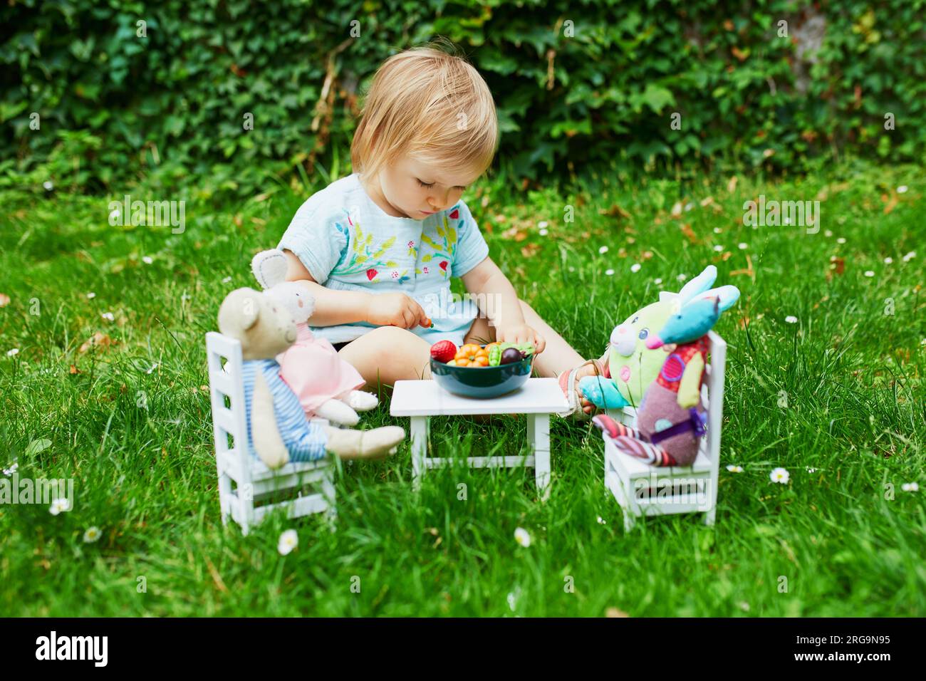 Adorable niña jugando con juguetes blandos en el parque o jardín y haciéndolos cenar con frutas y verduras de juguete. Juegos de rol para niños pequeños Foto de stock