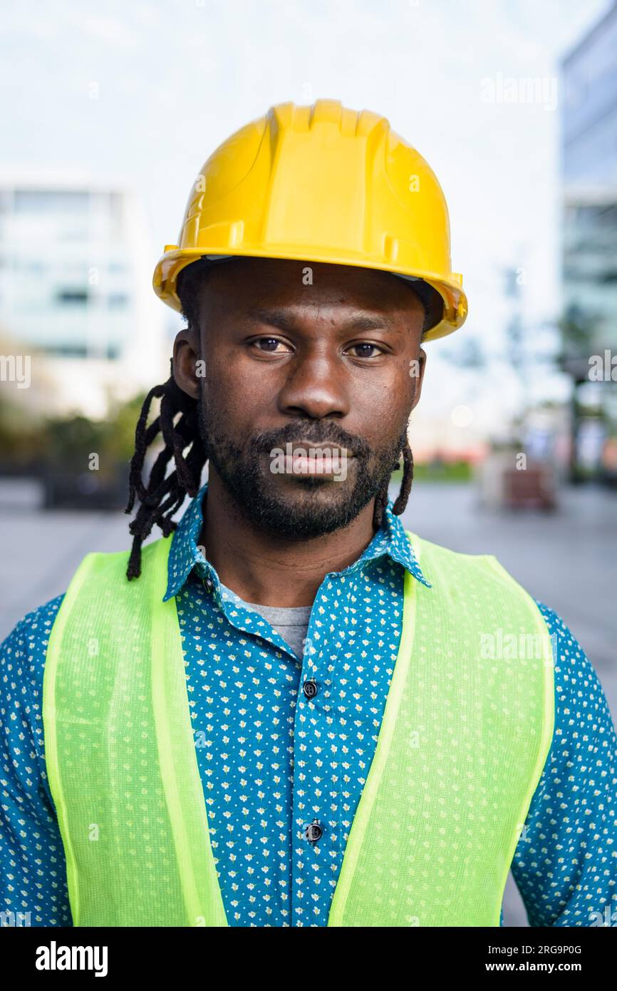 retrato vertical de hombre ingeniero negro con barba corta y rastas, que está usando casco y chaleco reflectante y está de pie al aire libre mirando Foto de stock