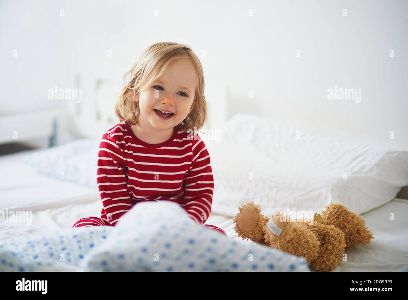 Niña feliz en pijama de rayas rojas y blancas sentado en la cama justo después de despertar. Siestas de día para niños pequeños Foto de stock