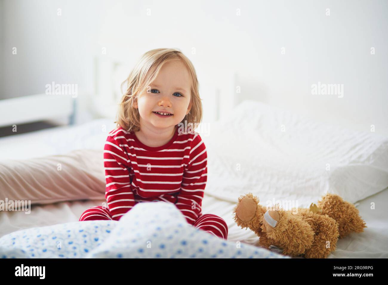 Niña feliz en pijama de rayas rojas y blancas sentado en la cama justo después de despertar. Siestas de día para niños pequeños Foto de stock