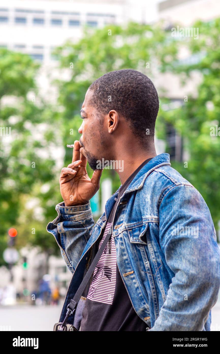 Hombre negro joven en chaqueta vaquera fumando cigarrillo - París, Francia. Foto de stock