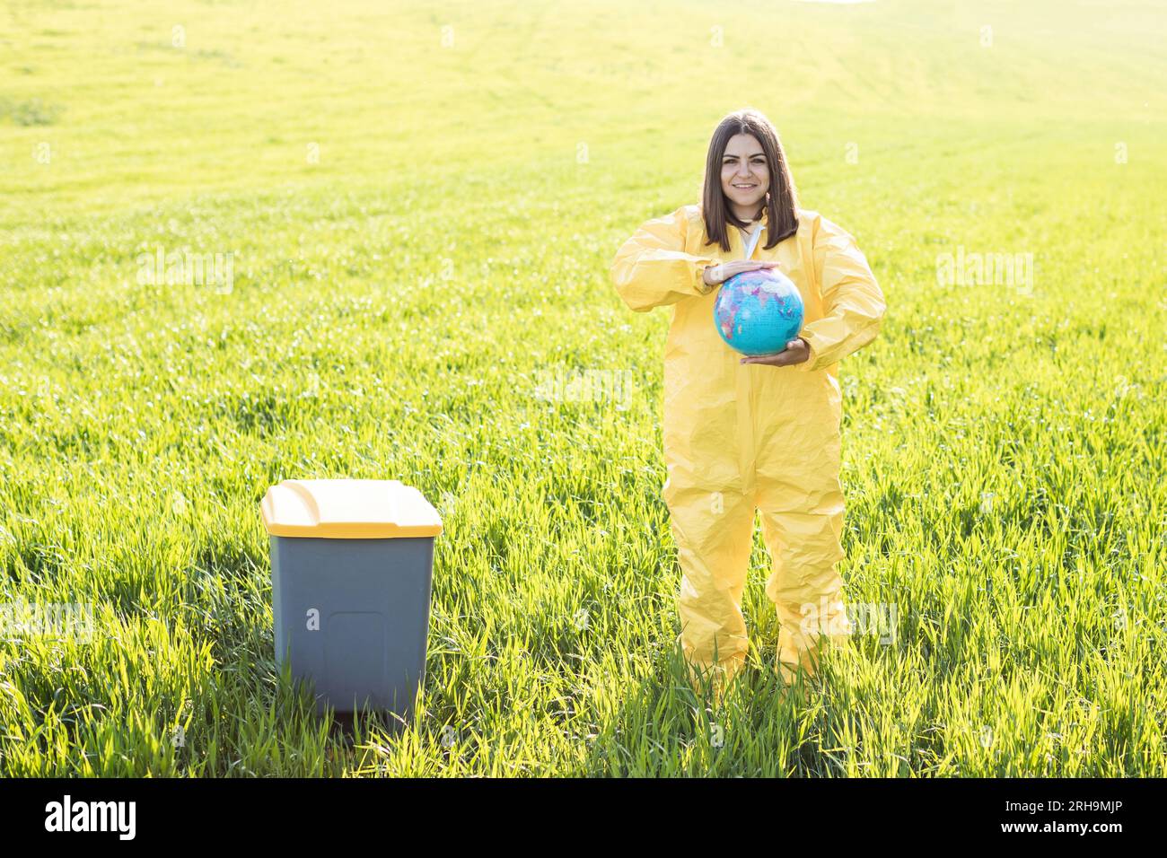 Una mujer en un traje protector amarillo está en medio de un campo verde y sostiene un globo en sus manos, junto a una papelera. El cuidado del planeta, excepto el Foto de stock